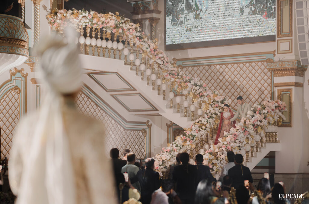 Indian bride walking down a flight of stairs decorated with pastel floral arrangements